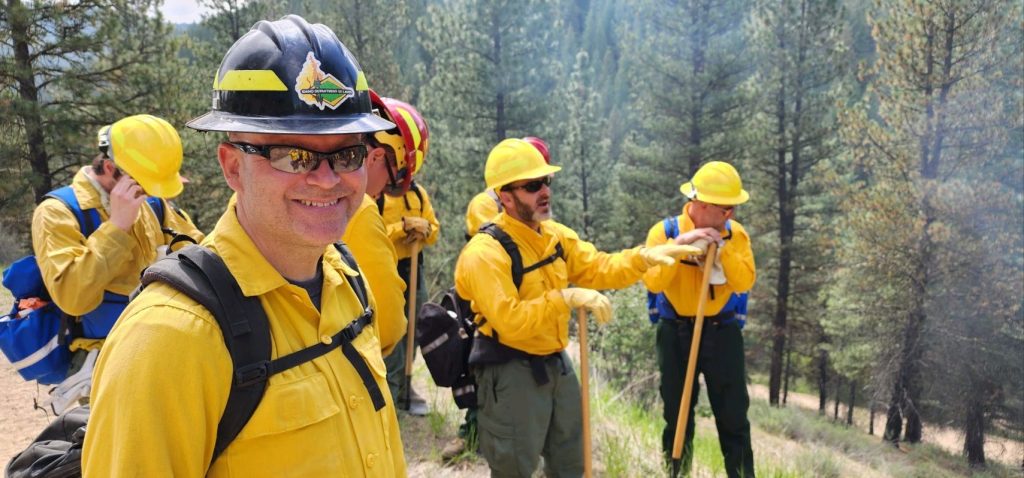 Idaho Secretary of State Phil McGrane wearing fire fighting equipment and standing with six other firefighters on a forest trail.